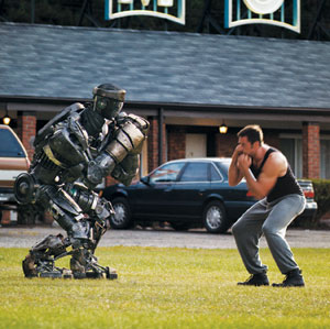 A man in workout clothes faces a large robot on a grassy area, both in boxing stances, with cars and a brick building in the background.