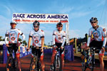 Four cyclists in matching uniforms pose with their bikes at the starting line under a banner reading Race Across America on a sunny day.
