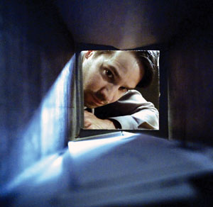 A man is looking through a narrow, rectangular opening, appearing to peer inside a dark tunnel or vent, with his face partially lit by light coming from behind him.