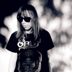 A person with long hair wearing dark sunglasses and a graphic t-shirt stands in front of a blurry, high-contrast background. The image is in black and white.