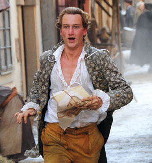 A young man in historical clothing runs outside on a cobblestone street, holding a wrapped package. He has blond hair and an open, patterned coat, with snow or frost on the ground behind him.