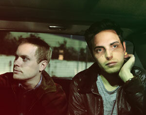 Two young men sit in the backseat of a car at night. One, wearing a brown jacket, looks out the window while the other, in a leather jacket, rests his chin on his hand and gazes ahead with a thoughtful expression.