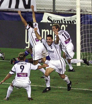 Four soccer players in white uniforms celebrate a goal on the field, raising their arms in excitement near the goalpost. One player wears number 9 and another wears number 10. A crowd and banners are visible in the background.