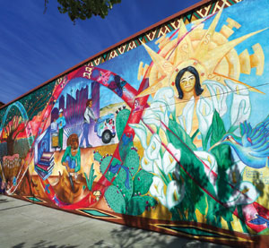 A vibrant mural on a building wall features colorful scenes including a woman surrounded by lilies, a cityscape, an ambulance, musical instruments, a cactus, and radiant sun imagery, celebrating community and culture.