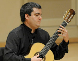 A person wearing a black shirt sits and plays a classical guitar, focusing intently on the instrument in a well-lit indoor setting.