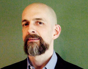 A bald man with a beard and mustache, wearing a blue collared shirt and dark blazer, stands in front of a plain green background, looking slightly to the side.