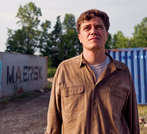 A man in a brown button-up shirt stands outdoors near a blue shipping container and a wall with graffiti, with trees and a partly cloudy sky in the background.