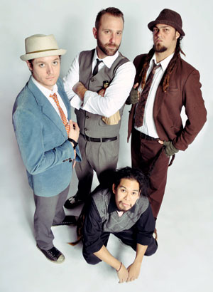 Four men pose together against a white background. Three stand wearing suits and hats, while one kneels in front, dressed more casually. They all look upward toward the camera with confident expressions.