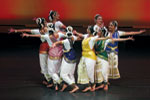 A group of dancers in colorful traditional costumes perform on stage with coordinated arm movements against a warm, gradient background.