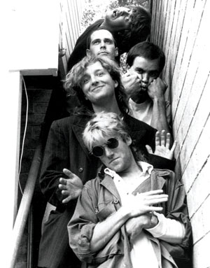 Five men stand closely together on an outdoor stairway between two walls, posing playfully. Some smile, one wears sunglasses, and another covers his mouth with his hands. The photo is black and white.