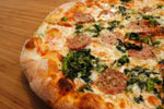 A close-up of a pizza on a wooden surface, topped with melted cheese, sausage slices, tomato sauce, and green leafy vegetables.