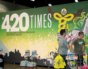 Two people stand by a booth with magazines and bags in front of a large display reading The 420 Times: The Magazine of Medical Marijuana and Natural Healing, with colorful cannabis-themed graphics in the background.