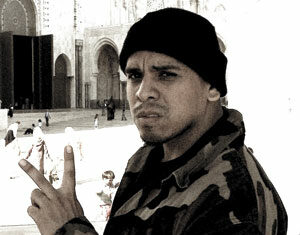 A man in a camouflage jacket and black beanie makes a peace sign with his fingers while looking seriously at the camera. He stands outdoors near a large building with arched doorways in the background.