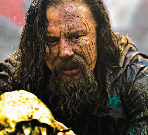 A man with long hair and a beard, his face smeared with dirt, looks intensely at a yellowed animal skull he is holding. He appears distressed and is wearing tattered, muddy clothing.
