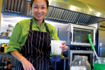 A person in a green shirt and striped apron smiles while holding a cup in a commercial kitchen, with stainless steel appliances and utensils visible in the background.