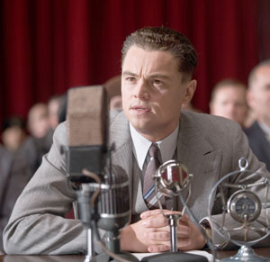 A man in a gray suit sits at a table with vintage microphones, speaking or listening intently, with a blurred audience and red curtains in the background.