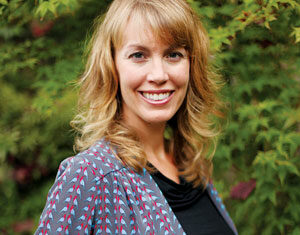 A woman with wavy blonde hair wearing a gray patterned jacket and black top smiles while standing outdoors in front of leafy green plants.