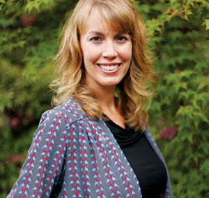 A woman with wavy blonde hair wearing a gray patterned jacket and black top smiles while standing outdoors in front of leafy green plants.