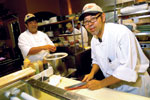 Two chefs in a restaurant kitchen preparing food; one chef is slicing fish on a cutting board while the other stands by a counter, both wearing white uniforms and caps.