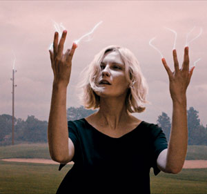 A person with short blonde hair stands outdoors, looking up with both hands raised. Lightning-like energy appears between their fingers, and the sky is cloudy in the background.