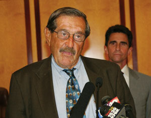An older man in a suit and tie speaks at a podium with microphones. A second man in a suit stands in the background. The setting appears to be an indoor press conference or formal event.