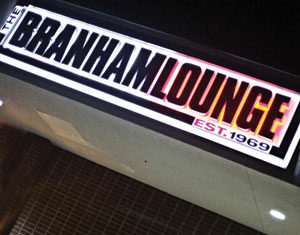 A neon sign reads THE BRANHAM LOUNGE EST. 1969 above a brick wall with the words Cocktails & Dreams in cursive, indicating a bar or lounge entrance at night.