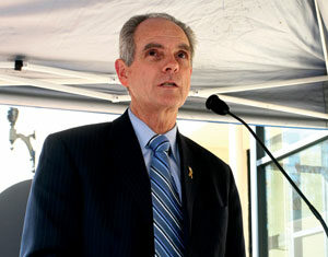 A man in a dark suit and striped tie speaks at a podium with a microphone, standing under a canopy, with a building and a streetlamp visible in the background.