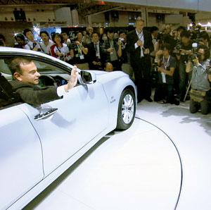 A man in a suit sits in a white car, waving through the open window, while a large crowd of photographers and onlookers gather around, taking pictures at an indoor event.