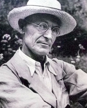 A black-and-white photo of an older man wearing round glasses and a straw hat, dressed in a collared shirt and jacket, looking slightly to the side with a thoughtful expression.