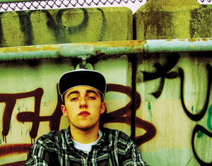 A young man in a plaid shirt and cap sits against a spray-painted, graffiti-covered wall, looking at the camera with a serious expression. A chain-link fence and concrete barrier are visible behind him.
