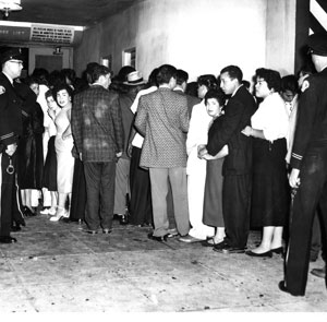 A black-and-white photo showing a group of people standing in line indoors, many facing away from the camera. Several women look toward the camera and two police officers stand at the edges of the scene.