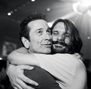 Two men embrace closely in a warm hug at a dimly lit event, both appearing happy and relaxed. One man smiles with his eyes closed while the other looks outward.