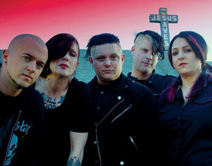 Five people dressed in dark clothing stand together in front of a building with a large cross in the background that reads Jesus Saves against a pink and blue sky.