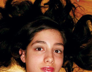 A young woman with long, dark hair lies on a wooden floor, her hair spread out around her head. She has a neutral expression and is wearing a colorful top.