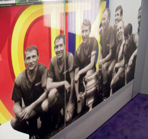A group of male soccer players in uniform sit and kneel together, smiling, with a large trophy in front of them. The background is colorful, featuring red, yellow, and blue designs.
