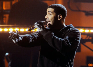 A man wearing black gloves and a black outfit holds a microphone and performs energetically on stage, with warm stage lights and blurred musical equipment in the background.