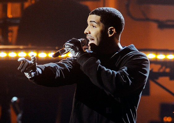A man wearing black gloves and a black outfit holds a microphone and performs energetically on stage, with warm stage lights and blurred musical equipment in the background.