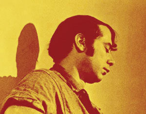 A man with dark hair plays an acoustic guitar, seated and facing left. The image has a yellow and brown color filter, and a shadow of the man is cast on the wall behind him.