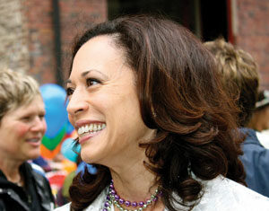 A woman with shoulder-length brown hair smiles while wearing a light-colored jacket, a white top, and beaded necklaces. She is outdoors with people and colorful balloons in the background.
