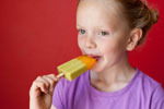 A young child in a purple shirt holds and eats a green and orange popsicle, standing in front of a bright red background.