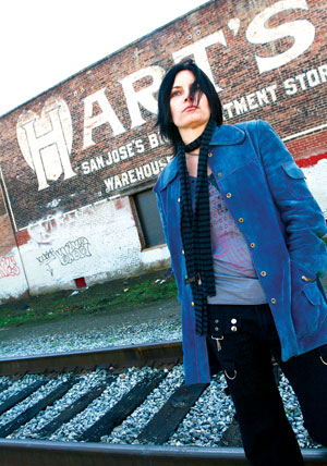 Person with long black hair wearing a blue coat and striped scarf stands near railroad tracks in front of a weathered brick building with a faded Harts warehouse sign.