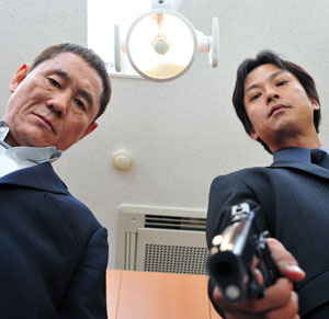 Two men in suits look down sternly, one pointing a handgun towards the camera. The image is shot from a low angle, with a ceiling light and vent visible above them.