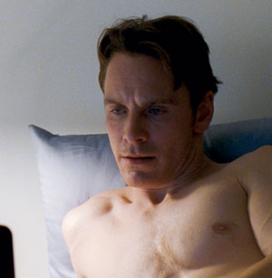 A shirtless man with light skin and short brown hair lies in bed, propped up by a pillow, looking intently at something off-screen in a softly lit room.