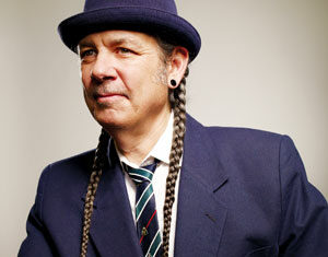 A person wearing a navy blue suit, striped tie, and navy hat with two long braided pigtails. They have stretched earlobes and are posing against a plain background, looking slightly to the side with a neutral expression.