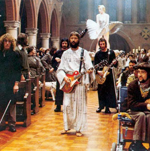 A group of musicians, including a man in a white robe playing guitar, walk down a church aisle filled with seated people; a woman in white and a flying dove are visible above the crowd.