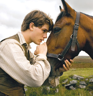 A young man gently holds a brown horse’s face, touching his forehead to the horse’s and looking at it with affection, outdoors in a grassy field under a cloudy sky.