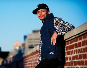 A man in a checkered shirt, dark vest, and white sneakers leans casually against a brick wall on a rooftop, with a cityscape blurred in the background and sunlight casting a warm glow.