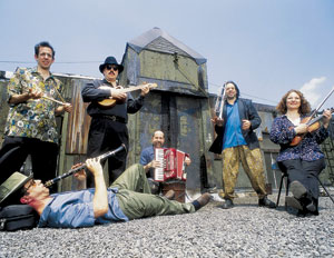 Five musicians with various instruments pose outdoors on a gravel surface near a building; one lies on the ground playing clarinet, while the others stand or sit holding string instruments and an accordion.