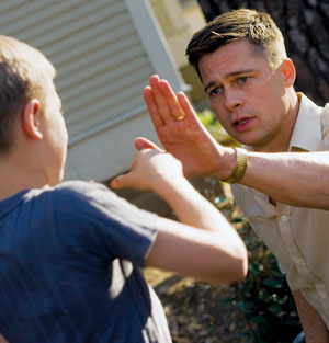 A man raises his hand while speaking to a boy outdoors. The man appears serious, and the boy faces him with his arms slightly raised. They are near a building and trees.