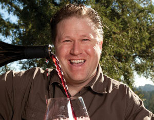 A smiling man outdoors pours red wine from a bottle into a glass, with trees and sunlight in the background.